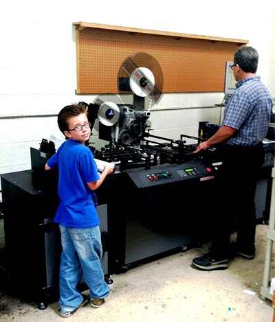 A man and a boy stand in front of a large machine. - sandiegodirectmail